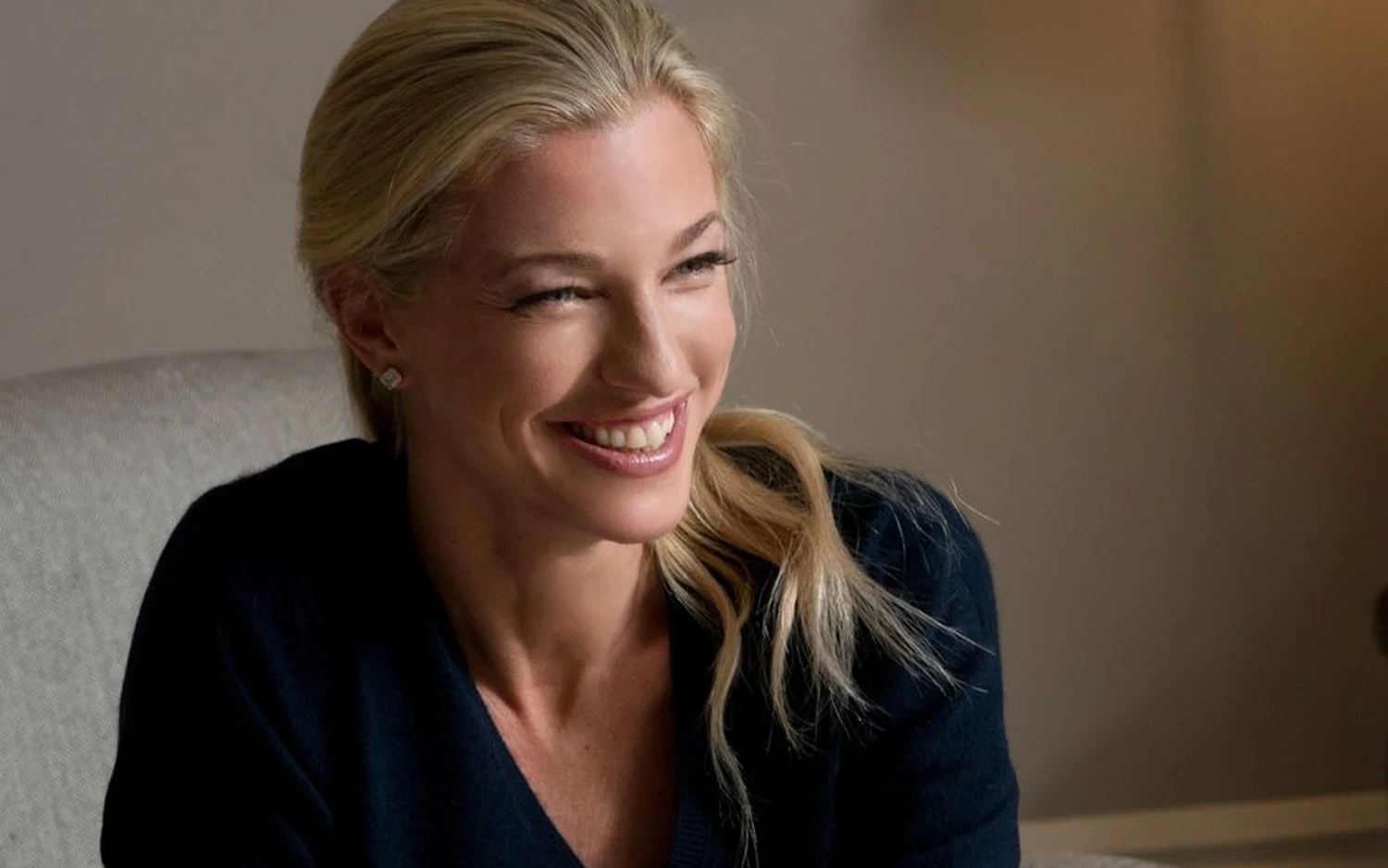 Nicole Junkermann seated in a pale armchair in a navy jumper, smiling to one side